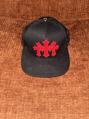 Chrome Hearts keps  - Snygg svart truckerkeps från Chrome Hearts med stort rött broderat motiv framtill och vit logga baktill. Kepsen har mesh på sidorna och baksidan för extra ventilation och en klassisk böjd skärm. Perfekt för en streetwear-look.