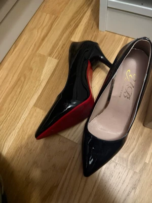 Svarta lackpumps med röd sula - Säljer ett par snygga svarta pumps från YGB i glansigt lackmaterial. Skorna har spetsig tå, klassisk klack och en cool röd sula som sticker ut. Perfekta för dig som vill ha en elegant och trendig look. Använda en gång.