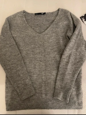 Grå stickad tröja med V-ringning - Mysig grå stickad tröja  med klassisk V-ringning och långa ärmar. Tröjan har ribbade muddar vid ärmslut och nederkant. Perfekt för lager på lager och enkel att matcha med olika stilar.
