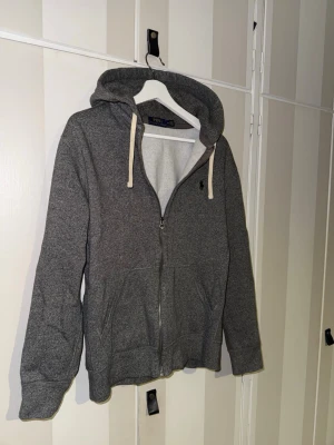 Grå hoodie från Polo Ralph Lauren - Snygg grå hoodie från Polo Ralph Lauren med dragkedja framtill och vita dragsnören i huvan. Klassisk logga broderad på bröstet och två fickor framtill. Tillverkad i mjukt sweatshirtmaterial som är perfekt för chill dagar.