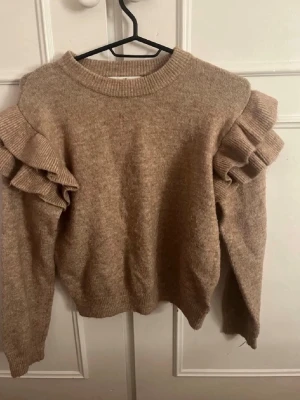 Beige stickad tröja med volangärm - Säljer en beige stickad tröja från H&M med unika volangdetaljer på axlarna. (Olika färg eftersom det är olika belysning med bild 2 stämmer). Finns ett litet hål