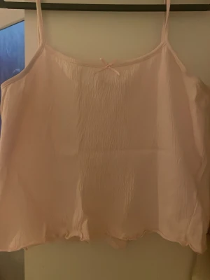 Ljusrosa linnetopp med rosett - Söt ljusrosa linnetopp med tunna axelband och liten rosett framtill. Toppen har en lätt vågig kant nedtill och är tillverkad i ett tunt, mjukt material som ger en luftig känsla. Använd ett fåtal gånger.