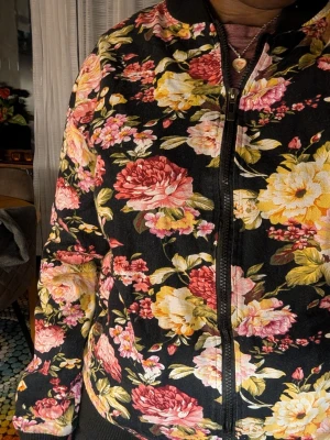 Blommig bomberjacka med dragkedja - Snygg bomberjacka med svart bas och stora blommor i rosa, gult och vitt. Jackan har dragkedja framtill, ribbade muddar och klassisk bomberkrage. Perfekt för dig som vill sticka ut med en färgglad och trendig look.