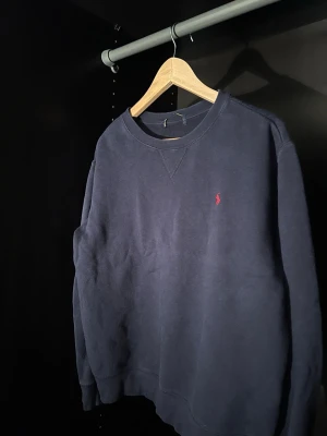 Mörkblå stickad tröja från Polo Ralph Lauren - Mörkblå stickad tröja från Polo Ralph Lauren med klassisk rund halsringning och den ikoniska röda logotypen broderad på bröstet. Tröjan har ribbade muddar vid ärmslut och nederkant, perfekt för en stilren och avslappnad look.