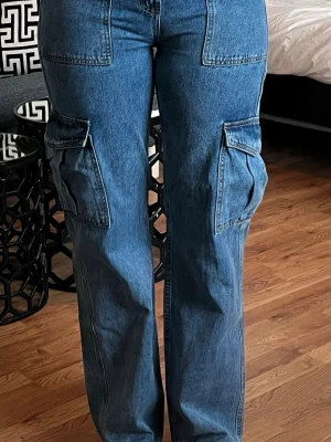 Blå cargobyxor i denim - Säljer ett par blå cargobyxor i klassisk denim med raka ben och stora fickor på sidorna. Byxorna har hög midja och tydliga sömmar. Storlek US 5 och jag är 173 cm. 