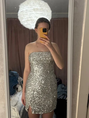 Glittrig silver kortklänning - Säljer en glittrig kort klänning i silver med paljetter över hela. Klänningen är axelbandslös och har en liten slits framtill. Perfekt för dig som vill sticka ut och glänsa på nästa fest. Materialet är polyester paljetterna ger ett riktigt partyvibe.