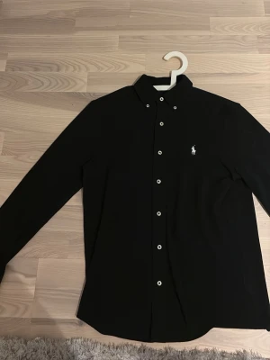 Svart skjorta från Polo Ralph Lauren - Svart långärmad skjorta från Polo Ralph Lauren med klassisk krage och vita knappar. Skjortan har broderad logga på bröstet och är tillverkad i ett lätt meshmaterial. Nypris 1699kr, storlek S.