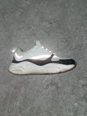  vita och svarta b22or - Säljer ett par chunky sneakers i vitt och svart med reflekterande detaljer. Skorna har en tjock sula och meshpartier för extra ventilation. Snygg kontrast mellan de svarta och vita partierna samt coola detaljer på ovandelen. Perfekta för dig som gillar streetwear och sportig stil.