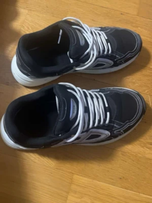 Svarta sneakers från Adidas - Snygga svarta sneakers från Adidas med vita detaljer och vita snören. Skorna har en sportig design med tjock vit sula och mönstrade sömmar. Perfekta för dig som gillar en klassisk streetwear-look.