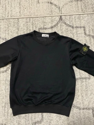 Svart sweatshirt från Stone Island - Snygg svart sweatshirt från Stone Island med klassisk rund hals och ribbade muddar. Tröjan har den ikoniska Stone Island-loggan på vänster ärm och är tillverkad i mjukt bomullsmaterial. Perfekt för en stilren och avslappnad look.