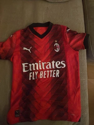 AC Milan matchtröja Puma röd/svart - Säljer en AC Milan matchtröja från Puma i rött och svart med grafiskt mönster. Tröjan har klubbmärke och Puma-logga på bröstet samt sponsortryck 'Emirates Fly Better' framtill. Klassisk V-ringning och korta ärmar. Perfekt för fotbollsfans!