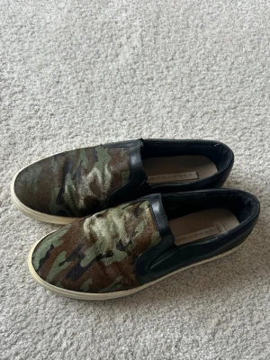 Camo slip-on sneakers med svarta detaljer - Säljer ett par slip-on sneakers med camouflagemönster i grönt, brunt och svart. Skorna har svarta läderdetaljer runt öppningen och vit sula. Perfekta för dig som gillar streetwear och vill sticka ut lite extra. Passar ca  37-38❤️
