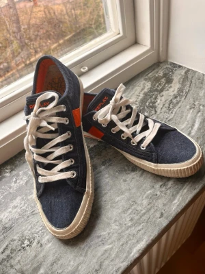 Replay denim sneakers med vita snören - Snygga låga sneakers från Replay i mörkblå denim med vita snören och gummisula. Skorna har orange detaljer och Replay-logga på plösen och sidan. Insidan är fodrad med orange tyg och sulan har ett rutmönster. Perfekta för en avslappnad streetstil.