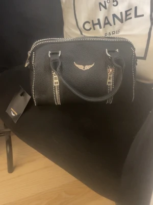 Svart handväska från Zadig & Voltaire - Snygg svart handväska i strukturerat läder från Zadig & Voltaire. Väskan har silverfärgade detaljer, dragkedjor och det ikoniska vingemblemet framtill. Två handtag och dekorativa nitar längs kanterna ger en cool och trendig look.