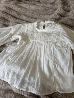 Vit romantisk blus med spetsdetaljer - Söt vit blus med långärmad design och fina spetsdetaljer. Blusen har en hög krage med volang och knäppning bak, samt ett luftigt och plisserat tyg som ger en bohemisk känsla. Perfekt för dig som gillar en romantisk och vintage-inspirerad stil.