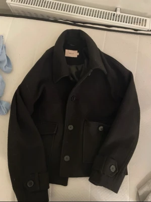Svart ulljacka från Nelly - Stilren svart jacka från Nelly i ullmaterial. Jackan har stora knappar framtill, två rejäla fickor och klassisk krage. Passformen är något loose och ärmarna är långa med knappdetaljer vid mudden. Perfekt för kyligare dagar.