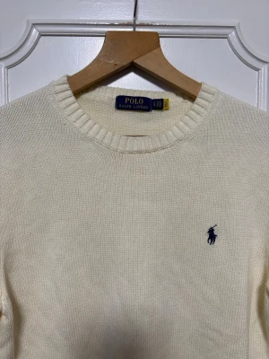 Vit stickad tröja från Polo Ralph Lauren - Klassisk vit stickad tröja från Polo Ralph Lauren med rund halsringning och ribbade muddar. Tröjan har den ikoniska blå broderade loggan på bröstet och är tillverkad i mjukt material som passar perfekt till lager på lager.