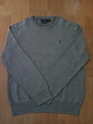 Grå stickad tröja från Polo Ralph Lauren - Klassisk grå stickad tröja från Polo Ralph Lauren med rund halsringning och diskret broderad logga på bröstet. Tröjan har ribbade muddar vid ärmslut och nederkant, perfekt för lager på lager under höst och vinter. Axelbredd: 46cm / Längd: 61cm. Nypris: 2295 / Mitt pris: 539kr