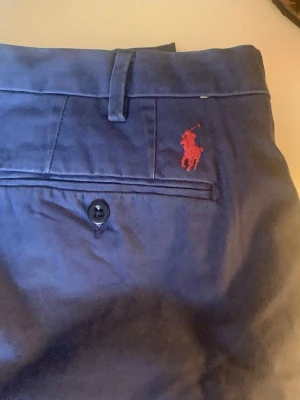 Blå chinos från Polo Ralph Lauren - Snygga blå chinos från Polo Ralph Lauren med röd broderad logga ovanför bakfickan. Byxorna har custom fit och är tillverkade i 100% bomull, vilket gör dem både bekväma och stilrena. Klassisk design med knapp på bakfickan och bälteshällor.