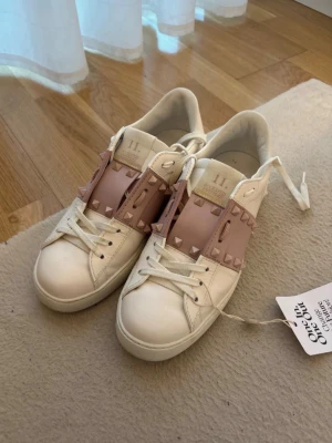 Valentino garavani sneakers - Snygga vita sneakers från Valentino med breda ljusrosa band över vristen dekorerade med ikoniska nitar. Skorna har klassisk låg profil, vita snören och detaljer i läder. Perfekta för dig som vill ha en unik touch på dina sneakers. beställda från Vestiaire och är äkta knappt använda 