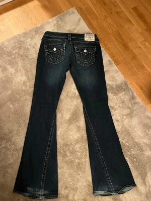Mörkblå bootcut jeans med fickdetaljer - Säljer mina True Religion jeans i modellen Joey (lågmidjade) Flare. Säljs på grund av att jag aldrig använder dem längre, och dom är lite för stora. Nere vid fötterna på baksidan är det lite små slitet, men det blir dom flesta långa jeansen, det är inget som syns framifrån. Köpte dom för 1300kr. Storlek 27 (M)