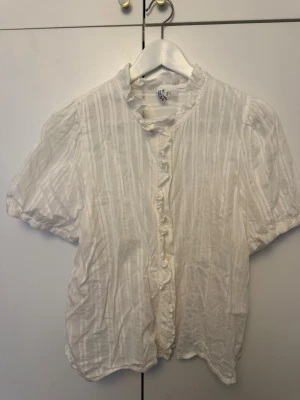 Vit blus med volang från H&M - Söt vit blus från hm med korta puffärmar och volangkant längs knäppningen. Blusen har diskreta ränder i tyget och en liten ståkrage med volang. Tillverkad i ett lätt och luftigt material, perfekt för en trendig och avslappnad look.