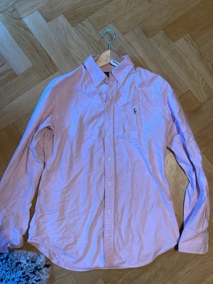 Ljusrosa skjorta från Ralph Lauren - Klassisk ljusrosa skjorta från Ralph Lauren med custom fit och ikonisk logga broderad på bröstet. Skjortan har långa ärmar, knappar framtill och en tidlös krage. Storlek medium men passar storlek s