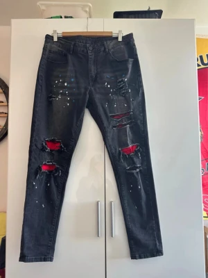 Svarta ripped jeans med röd detalj - Säljer ett par svarta slim fit jeans med stora slitningar och röda tygdetaljer under hålen. Jeansen har vita färgstänk och klassisk femficksmodell. Perfekta för dig som gillar en edgy och trendig stil.
