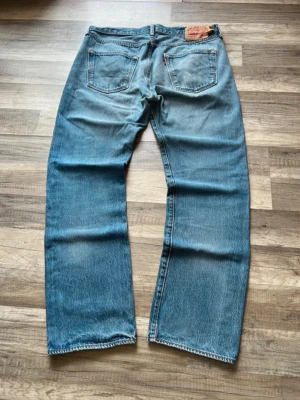 Levis 501 jeans med vintagekänsla - Säljer ett par klassiska blå jeans med rak passform och lätt slitna detaljer. Jeansen har fem fickor, normal midja och är tillverkade i denim. Perfekta för en avslappnad och tidlös stil.