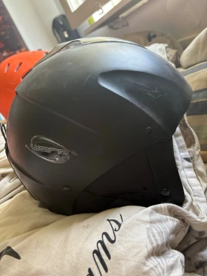 Svart hjälm från Warp - Säljer en svart hjälm från Warp med matt finish och öronskydd. Hjälmen har justerbart hakband och ventilationshål upptill. Perfekt för skidåkning, snowboard eller andra sporter där du vill skydda huvudet.