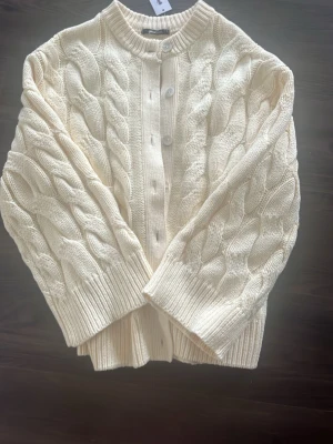 kabelstickad kofta Gina Tricot -  kabelstickad kofta i färgen vanilla yellow från Gina Tricot. Koftan har rund halsringning, stora stickade flätor, ribbade muddar och knäppning framtill med stora knappar. Helt ny men för stor! 