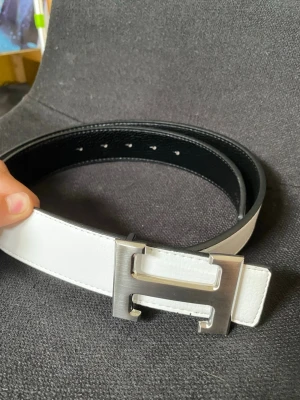 Vändbart vitt och svart skärp med H-spänne - Snyggt vändbart skärp i vitt och svart med ett stort metalliskt H-format spänne. Skärpet har en stilren design och passar perfekt till jeans eller kostymbyxor. Gjort i skinn för en exklusiv känsla.