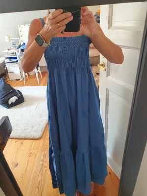 Blå långklänning med smock - Säljer en jeansklänning med smala axelband och smock upptill. Klänningen har en luftig passform och är perfekt för varma dagar. Tillverkad i ett mjukt tunnare jeans material och har en volang nedtill för extra flow.