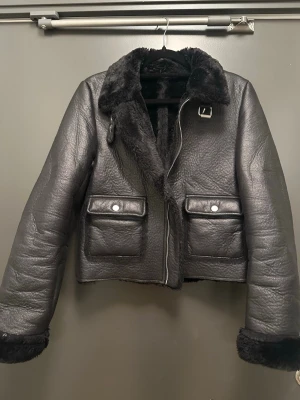 Svart bikerjacka med teddyfoder – fodrad skinnlook jacka - Snygg svart bikerjacka i skinnlook med mjukt teddyfoder och teddykrage. En trendig och tidlös jacka som håller dig varm under höst och vinter. Passar perfekt till jeans, kostymbyxor, kjol eller klänning och ger en stilren look. Har dragkedja, dekorativt spänne vid kragen och praktiska framfickor. 