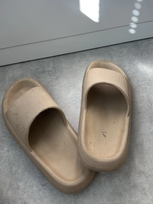 sliders/tofflor - Säljer ett par beige chunky tofflor med bred rem och upphöjd sula. Tillverkade i mjukt och flexibelt skummaterial som ger en bekväm känsla. Perfekta för att glida runt hemma eller på språng. Minimalistisk design utan synligt märke.