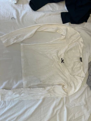 Vit långärmad topp med rosett - Söt vit långärmad topp med diskreta ränder och en liten svart rosett vid ringningen. Toppen har en croppad passform och är tillverkad i ett mjukt material som känns skönt mot huden. Perfekt för att skapa en clean och stilren look.