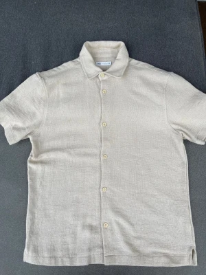 Beige linneskjorta från Zara - Säljer en stilren beige skjorta från Zara i linne. Skjortan har korta ärmar, klassisk krage och knappar framtill. Perfekt för dig som gillar en avslappnad men ändå snygg look. Passar till både jeans och shorts.