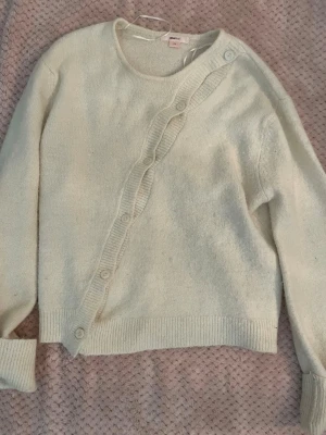 Vit kofta från Gina Tricot - Mysig vit kofta från Gina Tricot med rund halsringning och dekorativa knappar i sned rad framtill. Koftan har långa ärmar och ribbade muddar vid ärmslut och nederkant. Perfekt för lager på lager och enkel att styla till olika outfits.