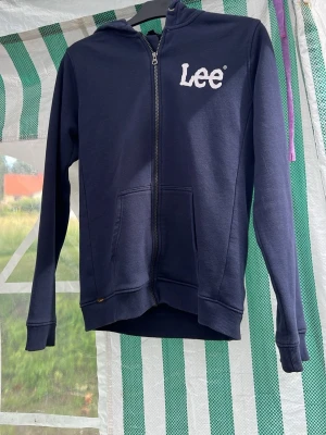 Mörkblå zip hoodie från Lee - Snygg mörkblå hoodie från Lee med dragkedja framtill och vit logga på bröstet. Regular fit-modell med huva och två fickor fram. Perfekt för en avslappnad och cool stil. Tillverkad i mjukt bomullsmaterial för extra komfort.