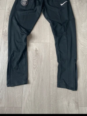 Svarta Nike PSG träningsbyxor - Säljer ett par svarta träningsbyxor från Nike med PSG-logga på ena benet. Byxorna har dragkedjor vid bensluten, elastisk midja och fickor. Materialet är lätt och andas, perfekt för träning eller chill. Swoosh-logga i vitt på framsidan.