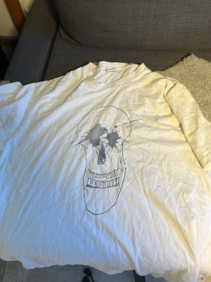 Vit t-shirt med dödskalletryck från Supreme - Vit t-shirt i bomull från Supreme med ett stort svart dödskalletryck på framsidan. Klassisk rund hals och korta ärmar. Perfekt för dig som gillar streetwear och schyssta plagg. Dock fanns det ett väldigt litet hål i armvecket som jag har sytt ihop. Hör gärna av dig vid funderingar eller prisförslag!!
