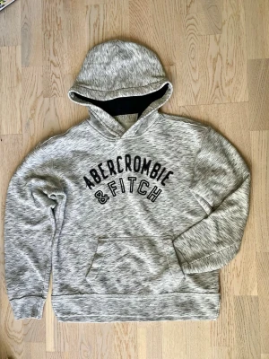 Grå hoodie Abercrombie & Fitch - Snygg gråmelerad hoodie från Abercrombie & Fitch med stor svart logga på bröstet. Hoodien har en klassisk huva och en stor magficka framtill. Mjuk och skön bomullsblandning, perfekt för chill dagar. Passar dig som gillar enkel men stilren streetwear. Funkar bra på tjejer och killar. Små skavanker som fläckar kan ses vid noga inspektion (se bild 3).