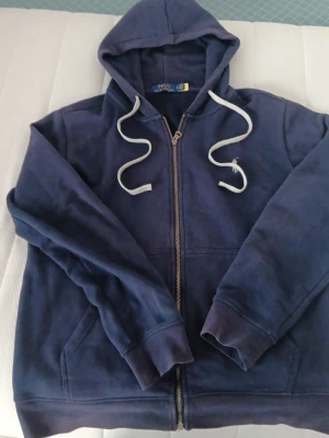 Marinblå hoodie från Polo Ralph Lauren - Snygg marinblå hoodie från Polo Ralph Lauren med dragkedja framtill, huva med vita snören och ribbade muddar. Tillverkad i mjukt bomullsmaterial som känns skönt mot huden. Perfekt för en avslappnad och stilren look.