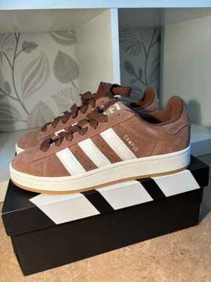 Bruna Adidas Campus sneakers - Helt nya och oanvända bruna Adidas Campus sneakers i mocka med klassiska vita ränder på sidorna och vit sula. Skorna har bruna skosnören och gummisula med mönster undertill. Perfekta för en avslappnad streetwear-look.