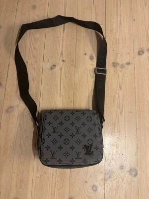 Grå axelväska med monogram - Snygg grå axelväska  med klassiskt svart monogrammönster och justerbar axelrem. Väskan har en fyrkantig form och är tillverkad i ett tåligt material. Perfekt för dig som vill ha en trendig praktisk väska.