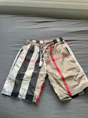 Beige-randiga shorts med snörning - Snygga shorts med breda ränder i beige, svart, vitt och röda detaljer. De har elastisk midja med vit snörning och avslappnad passform. Perfekta för sommaren och har en modern streetwear-vibe.