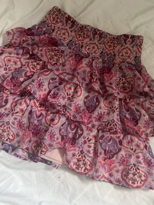 Rosa volangkjol från Lindex - Sitter som storlek xs. Supersöt kort volangkjol från Lindex i flera nyanser av rosa och lila med blommigt mönster. Kjolen har bred smock i midjan och flera lager volanger som ger en lekfull look. Perfekt för dig som gillar färg och mönster!