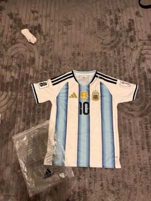 Argentina Messi matchtröja Adidas - Säljer en officiell Argentina matchtröja från Adidas med Messi och nummer 10 på ryggen. Tröjan har klassiska blå och vita ränder, svarta detaljer på axlarna och flera broderade märken på bröstet. Perfekt för fotbollsfans som vill stötta sitt lag.