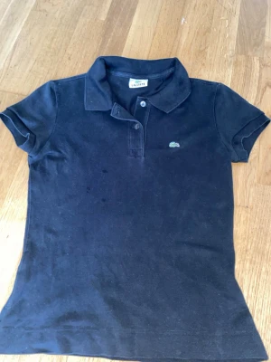 Svart pikétröja från Lacoste - Klassisk svart pikétröja från Lacoste med korta ärmar och krage. Tröjan har knappar framtill och den ikoniska gröna krokodilloggan broderad på bröstet. Tillverkad i bomull för en bekväm känsla och snygg passform.