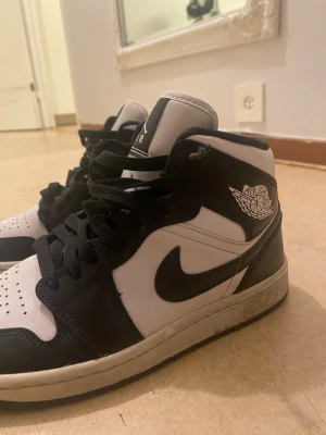 Svarta och vita Air Jordan 1 sneakers - Säljer ett par klassiska Air Jordan 1 sneakers i svart och vitt. Skorna har hög modell, svart swoosh och vingar-logga på sidan. Ovandelen är i läder med perforerad tåbox och svarta skosnören. Perfekta för dig som gillar streetwear och ikoniska sneakers.